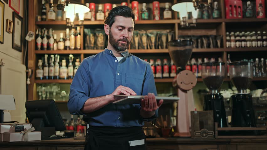 Bartender standing by cafe counter and opening laptop for checking online orders in application. Concentrated man using modern gadget for efficient work and quick communication with customers.