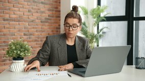 Young businesswoman in glasses and a blazer, working in an office. She conducts a video conference through her laptop, showing various charts related to business reporting. Camera 8K RAW