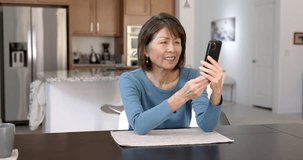 Cheerful attractive woman smiling enjoying video chat laughing talking using mobile phone relaxing at home table. Mature asian beauty streaming interesting internet blogger communication.
