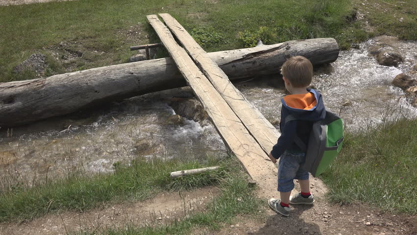 Little hiker with backpack crossing a wooden bridge over the mountain creek