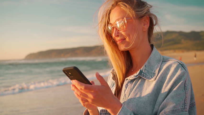 Smiling young woman with blond hair using smartphone on beach. Beautiful female in eyeglasses and denim shirt watching video on mobile phone. Lady enjoys her summer vacation on ocean shore.
