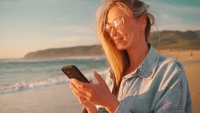 Smiling young woman with blond hair using smartphone on beach. Beautiful female in eyeglasses and denim shirt watching video on mobile phone. Lady enjoys her summer vacation on ocean shore. - Powered by Shutterstock - Get 15% off with code: PIKWIZARD15