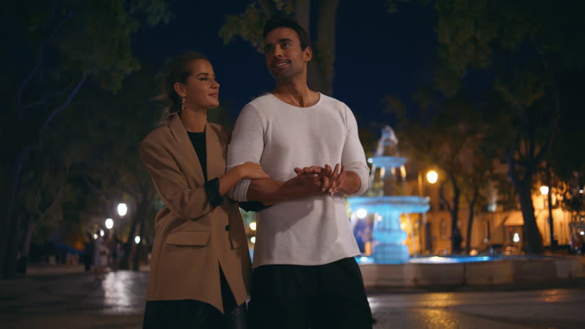 Romantic spouses walking city at night. Attractive muscular man talking embracing girl on evening stroll. Relaxed smiling couple family going park together. Beautiful loving woman holding guy hand.