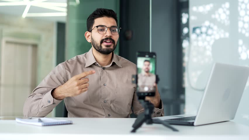 Confident bearded businessman in a shirt private tutor giving video call lesson or training using smartphone sitting at workplace in business office. Blogger is streaming. Coach has an online course