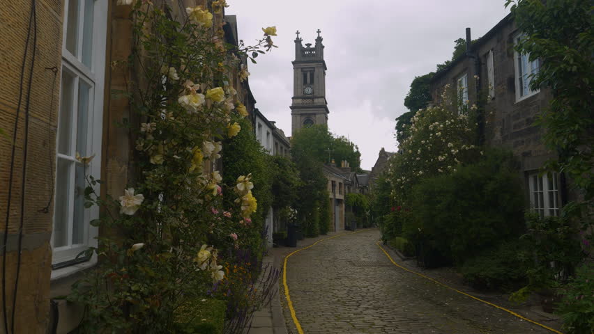 Charming street with old stone townhouses lined with blooming roses and lush greenery. Church tower rising above the picturesque winding cobbled alley near historic center of Scottish capital city.
