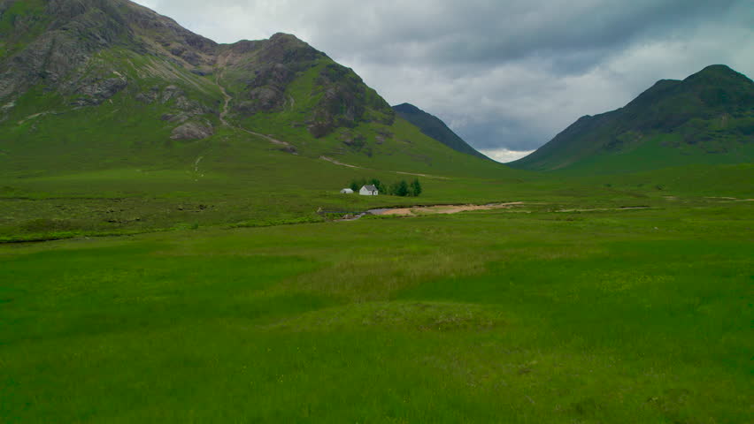 AERIAL: Small cottage by a winding river in the middle of a green grassy valley. Remote white hut below majestic mountain peaks and dark clouds rolling above dramatic landscape in Scottish Highlands.