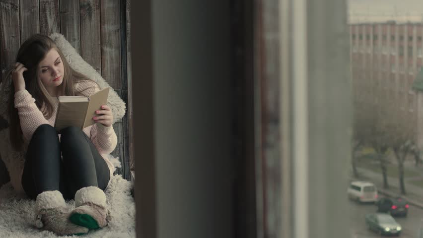 Beautiful young woman sitting on window and learn. Blond model girl read a book turning pages of the book holding hands close up. Natural lighting indoors