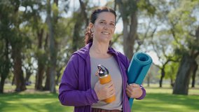 Positive mature woman in sports outfit holding exercise mat and water bottle, smiling and posing for camera after fitness class in the park. Medium shot, video portrait