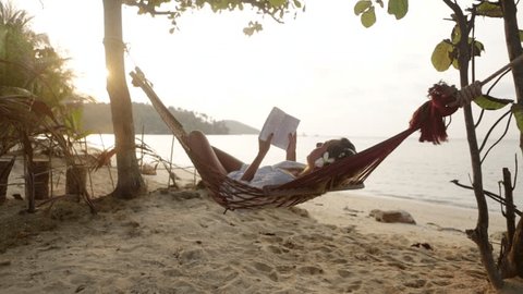 Young woman reading a book while lying down on a hammock surrounded by nature. Woman on hammock reading a book by the beach at sunset, female relaxing on hammock by the beach 