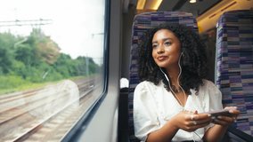Front view of young businesswoman commuting sitting by window on moving train streaming film or show to mobile phone wearing earphones looking out of window - shot in slow motion - Powered by Shutterstock - Get 15% off with code: PIKWIZARD15