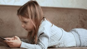 Cute girl with smartphone lying on sofa . Side view of child girl hands using mobile phone in the living room at home, typing message, 4k slow motion