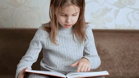 Child girl reading a book sitting on a couch, 4k slow motion. Cute girl reading a book sitting on sofa indoors, front view
