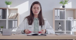 Portrait of smiling friendly female doctor cardiologist sitting at the desk on workplace holding red heart in hands and looking at camera in medical office. Medicine and cardiology concept. 4K video.