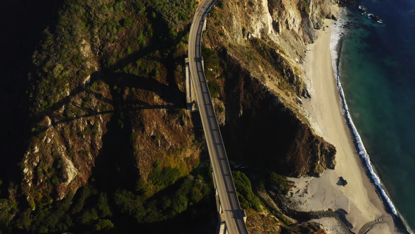 4k aerial footage of Bixby Creek Bridge on the Big Sur coast of California. Ocean waves at sunset by a rocky coast. A white car drives on a bridge along the coast. Road to the San Francisco  Royalty-Free Stock Footage #3414319361