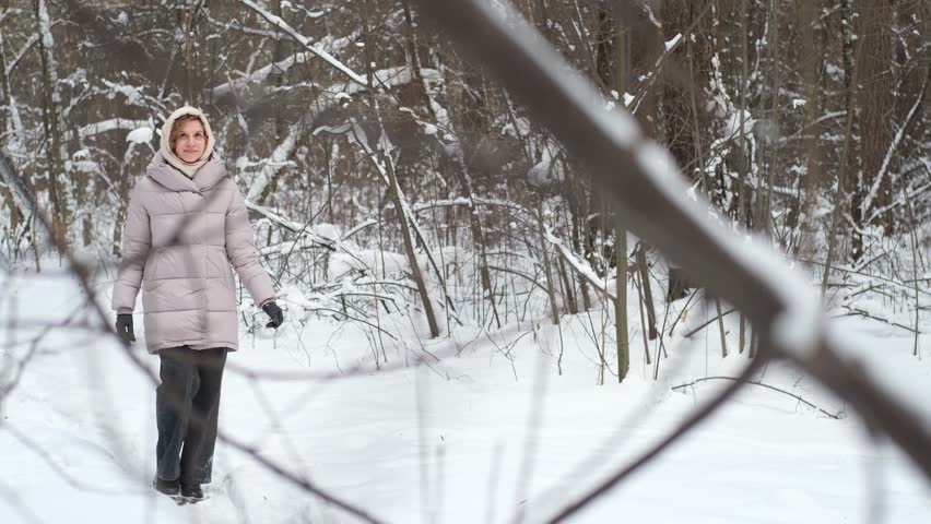 A young woman stands against a background of fir trees under a snowy forest in winter. A girl in a gray down jacket with a hood stands and looks at the camera. Close-up.