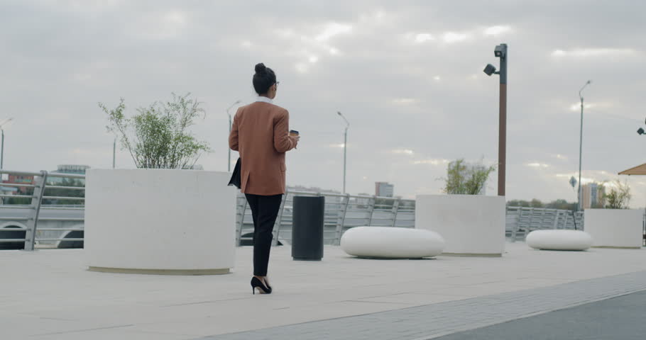 Slow motion of beautiful young woman walking along river bank in city center and drinking to go coffee. Metropolis and modern urban lifestyle concpt.