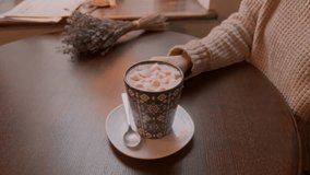 Close-up of a girl taking a cup of cocoa with marshmallows at the table. A cup with cocoa and marshmallows and the girl's hands are in the frame. The video was shot in 4k quality