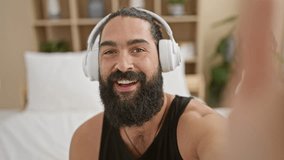 A smiling bearded young man wearing headphones enjoys a video call at his cozy bedroom.