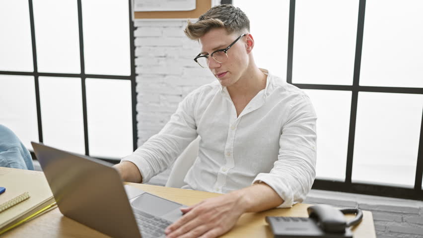 Happy and confident, young caucasian man working on his laptop in an indoor office workplace. business worker nailing his job with unwavering confidence and a beaming smile.