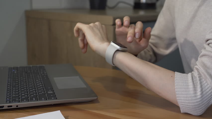 Girl is sitting at the kitchen table, typing text on a laptop and answering the call of a smartwatch