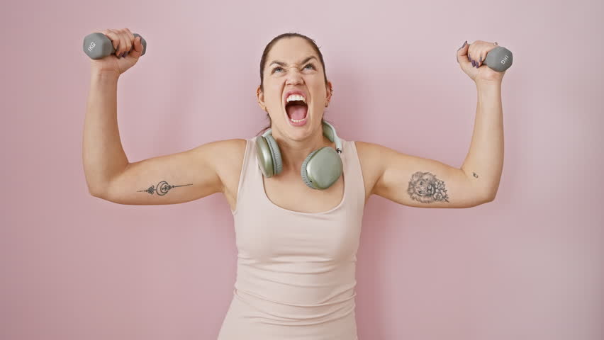 Furious young woman in sportswear, blue eyes ablaze in anger, shouting enraged with headphones on and weights in hand, expressing mad frustration over isolated pink background