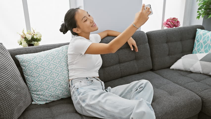 A young asian woman takes a selfie while sitting casually on a grey couch indoors, exuding happiness and comfort.