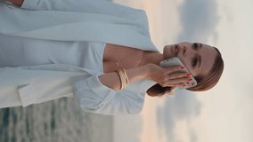Businesswoman speaking smartphone at sea sunset wearing white elegant suit vertical shot close up. Confident serious lady finish phone call standing at beautiful sundown sky alone. Busy woman hang up