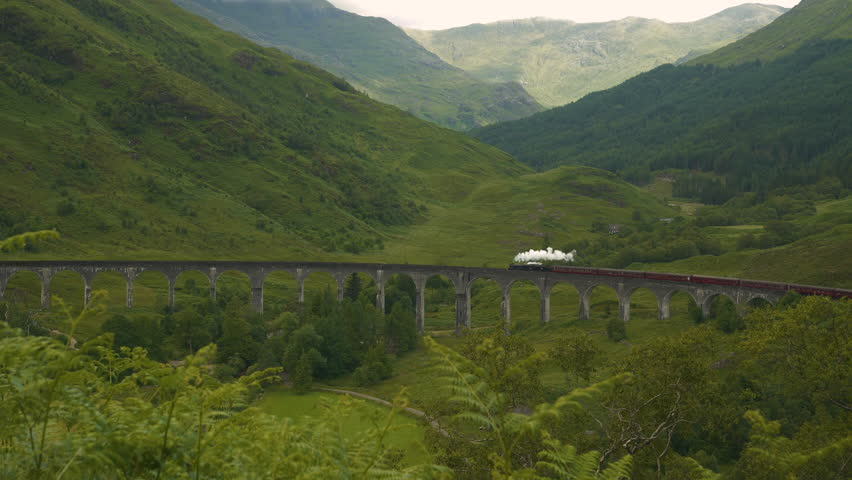 Picturesque stone viaduct over green valley crossed by a Jacobite steam train. Old locomotive on a railway journey through idyllic landscape of the Scottish Highlands with green valleys and mountains.