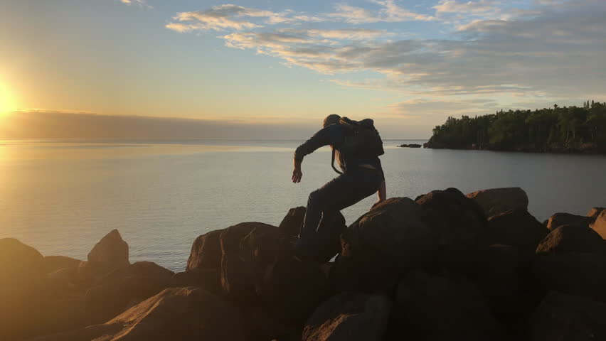 Ocean, rock and man hiking at sunset outdoor in nature, freedom and travel. Sea, adventure and person trekking in environment on summer holiday, relax on vacation and view by beach water on journey