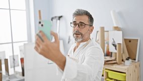 Handsome young hispanic, grey-haired male artist engrossed in a serious video call at indoor art studio, painting with paintbrush on canvas, learning and discussing art lessons.