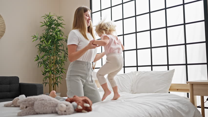 Caucasian mother and daughter bonding, jumping on the bed together, smiling inside their cozy bedroom, waking up a happy home with joyful expressions. Royalty-Free Stock Footage #3415033921