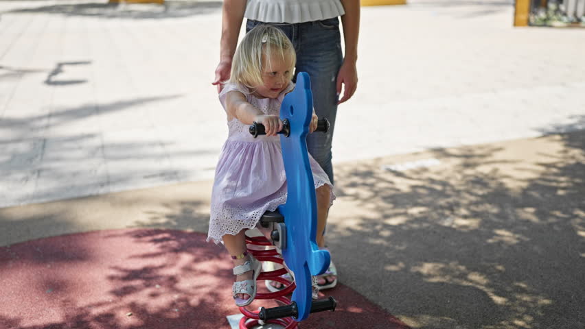 Caucasian mother and willing daughter sitting on a seesaw, playing seriously in the park playground enjoying family time outdoors