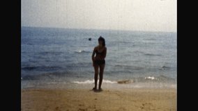 Woman in swimsuit walk along sandy shore on blue sea background. Adult happy woman in enjoying sunny beach holiday in warm country. Hot ocean vacation. Vintage color film. Family archive. Retro 1980s