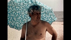 Funny man in swimsuit walking with sun umbrella on sandy beach. Man with humor, joking, laughing on sea shore. Cheerful vacationer on resort. Vintage color film. Holiday archive. Retro vacation 1980s