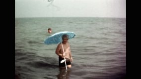 Man have fun in sea water under sun umbrella. Funny smiling adult vacationer joking, swimming in ocean. Cheerful man on beach vacation. Humor in summer holiday. Archival old film. Retro archive. 1980s