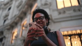 Cheerful African-American woman in 20s scrolling on smartphone, with headphones, against a historic building at twilight. She’s in a sleeveless top and glasses, embodying a tech-savvy urban charm