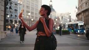Tech-savvy female enhances stories with playful filters, sharing city stroll through social media app. Smiling African American woman enjoy video call with frontal smartphone camera during travel