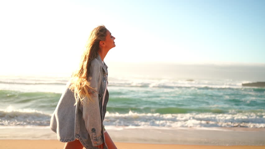 Portrait of smiling blond woman standing arms outstretched on sandy beach. Beautiful female in denim jacket and shorts standing near big rock and sea waves. Lady enjoys summer vacation. Royalty-Free Stock Footage #3415298637
