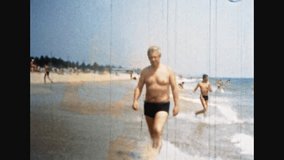 Man in swimsuit walking along sea shore. Relaxing hispanic person on people crowd backdrop. Ocean summer vacation on popular beach resort. Eighties memories. Vintage color film. Retro archive. 1980s