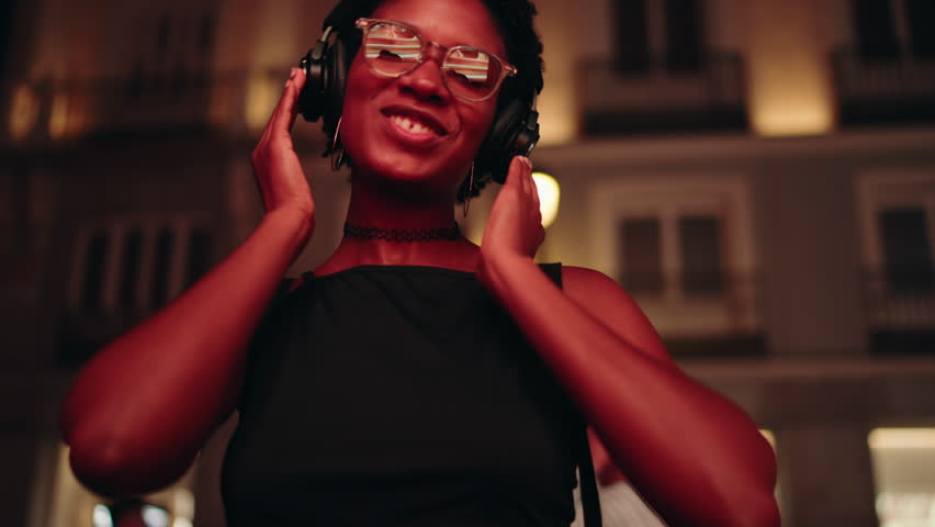 Excited young woman dances on night city, her headphones delivering perfect beat, she moves with sincere joy. Lost in melody, female enjoy listening online music celebrating good mood. Slow motion