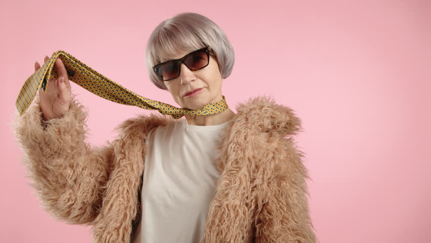 A woman in a fur coat with a necktie elegantly poses in front of the camera, playfully holding the tie in her hand. She has short blonde hair and wears sunglasses. The background is pink. 8K RAW.