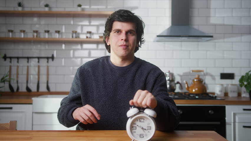 Young man holding alarm clock, showing time deadline, gestures no and shakes his head while sitting in the kitchen