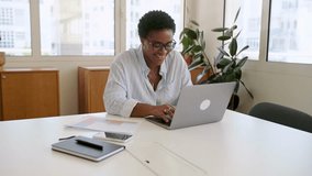 Side view on African-American businesswoman using laptop sitting at the desk at office, smiling charismatic female employee, freelancer wearing casual blue shirt and glasses working - Powered by Shutterstock - Get 15% off with code: PIKWIZARD15