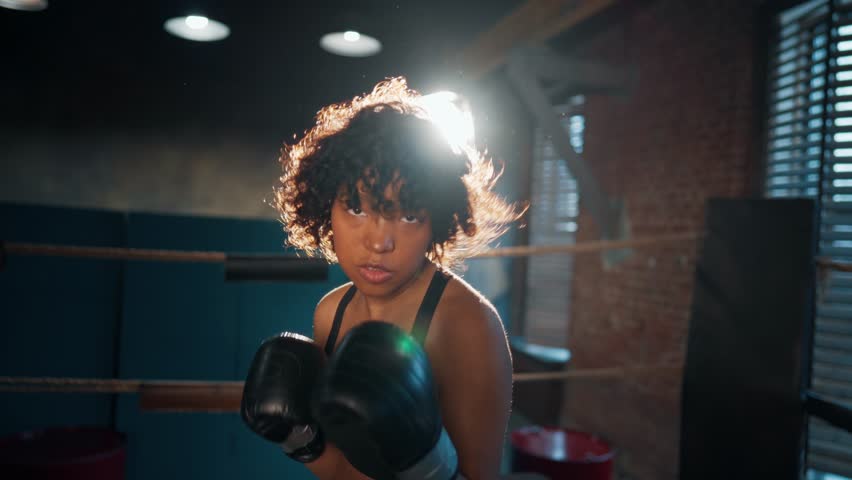 African american woman boxer stands on boxing ring and boxing. Sportswoman training in gym and looks at camera. Girl's power, woman train workout, sport fight, self-defense and feminism.