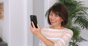Happy attractive woman smiling enjoying face time video chat talking using mobile phone relaxing standing at home. Mature asian beauty streaming internet blogger for communication palm background.