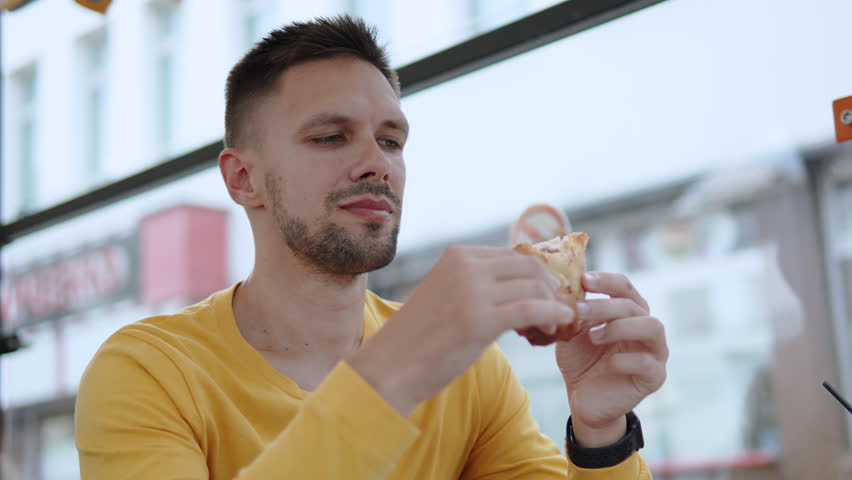 Portrait of slender, handsome brunette eating fried flatbread on terrace in sidewalk cafe. Man eats fast food snack, chewing food thoroughly, slow motion. Rapid restoration of body energy balance.