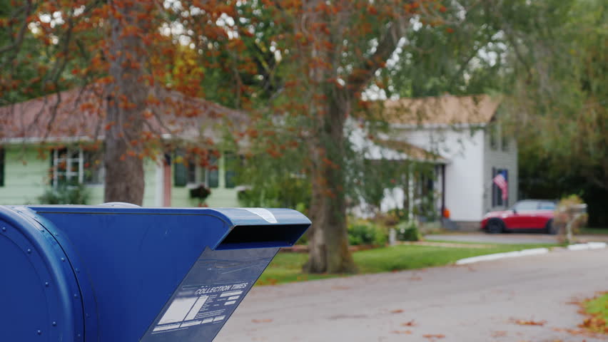 A woman with a dog in her arms approaches a mail box on the street and throws a letter into it