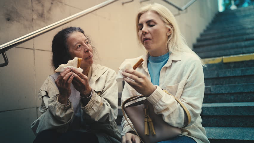 Pleased old female refugee and kind woman eating sandwiches together, charity