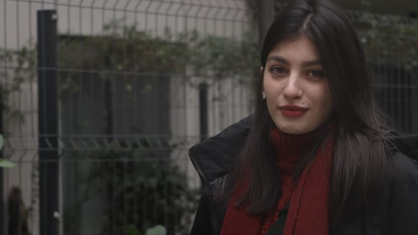 Elegant young brunette with red lips following the camera with her eyes while walking down the houses walkway.
