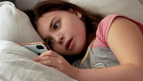 A little girl is sick, lying in bed weak and sad, looking into the frame in pajamas with disheveled hair, watching a video on a smartphone.